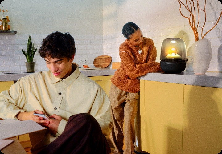 식탁에 기대어 앉은 남성은 휴대폰을 하고 있으며, 그 뒤에는 주방 서랍장에 기댄 여성이 노란색 불빛이 들어온 스피커를 바라보고 있습니다.
