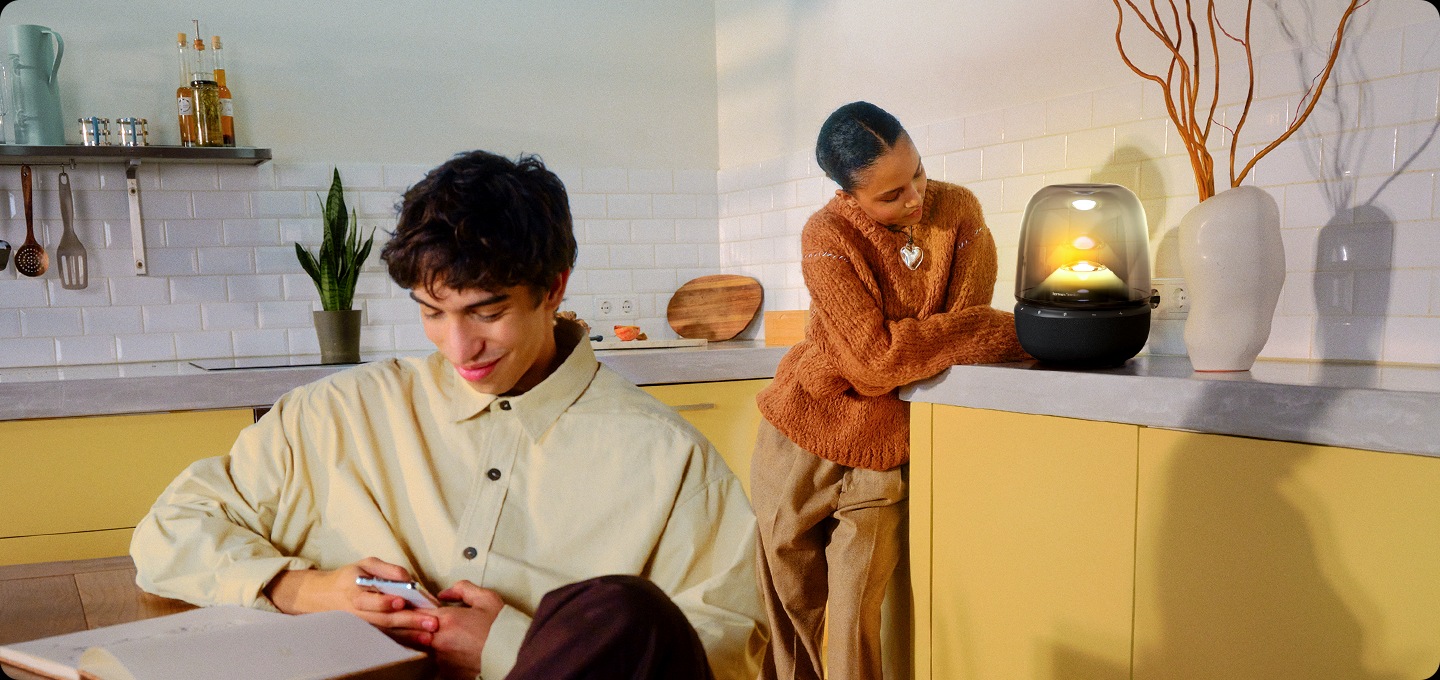 식탁에 기대어 앉은 남성은 휴대폰을 하고 있으며, 그 뒤에는 주방 서랍장에 기댄 여성이 노란색 불빛이 들어온 스피커를 바라보고 있습니다.