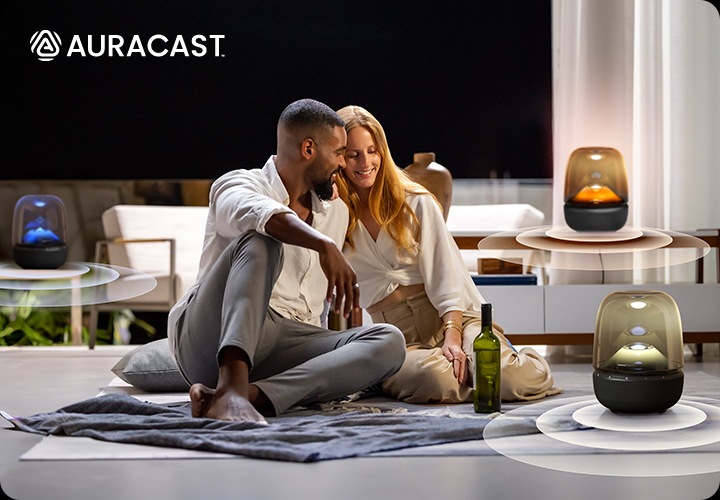 남성과 여성이 서로 기대어 거실에 앉아 있습니다. 남성과 여성 앞에는 와인 병과 노란색 불빛이 들어온 스피커가 놓여 있으며, 뒤에는 각각 테이블과 장식장 위에 파란색과 주황색 불빛이 들어온 스피커가 놓여 있습니다.