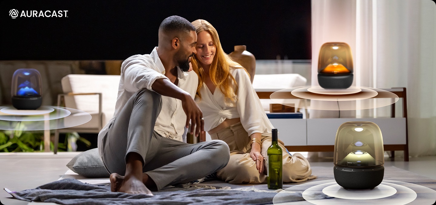 남성과 여성이 서로 기대어 거실에 앉아 있습니다. 남성과 여성 앞에는 와인 병과 노란색 불빛이 들어온 스피커가 놓여 있으며, 뒤에는 각각 테이블과 장식장 위에 파란색과 주황색 불빛이 들어온 스피커가 놓여 있습니다.