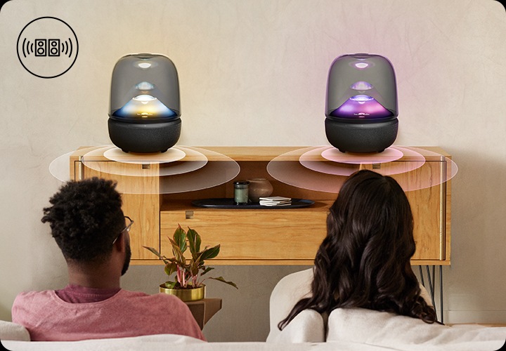 남성과 여성이 소파에 앉아서 장식장 위에 놓인 스피커들을 보고 있습니다. 스피커에는 파란색과 노란색 불빛이 나오는 스피커와 보라색과 분홍색 불빛이 나오는 스피커가 놓여 있습니다.