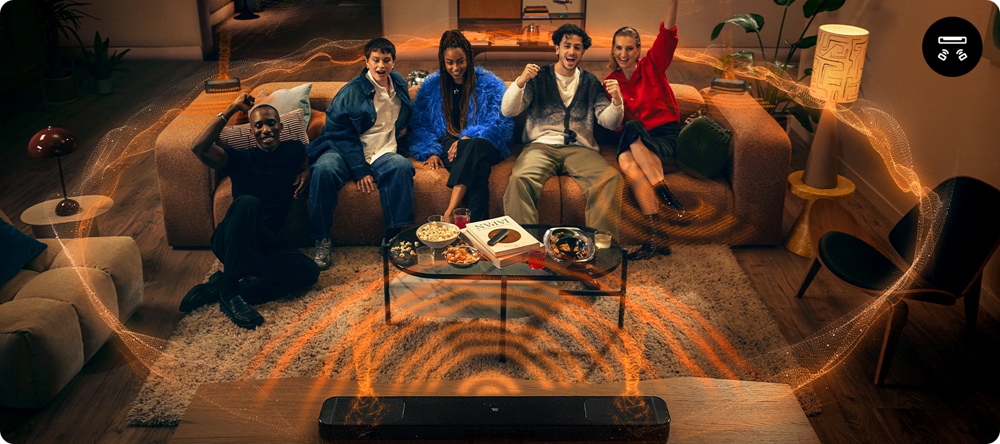 여성 2명과 남성 2명은 소파위에 앉아 있으며, 남성 1명은 소파에 기대어 앉아 있습니다. 사람들이 바라보는 곳에는 장식장 위에 사운드바가 놓여 있습니다. 사람들이 앉은 소파 양 끝에는 리어스피커가 놓여 있으며, 사운드바와 리어스피커에서 음향효과가 나오고 있습니다.