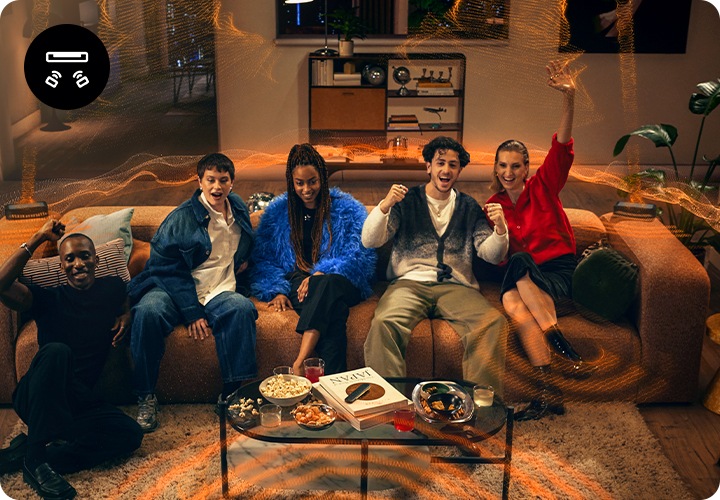 여성 2명과 남성 2명은 소파위에 앉아 있으며, 남성 1명은 소파에 기대어 앉아 있습니다. 사람들이 바라보는 곳에는 장식장 위에 사운드바가 놓여 있습니다. 사람들이 앉은 소파 양 끝에는 리어스피커가 놓여 있으며, 사운드바와 리어스피커에서 음향효과가 나오고 있습니다.