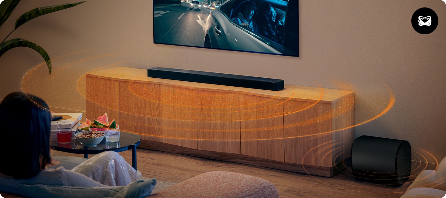 여성이 소파에 앉아서 TV를 보고 있습니다. TV 아래에는 장식장이 놓여 있으며, 장식장 위에는 사운드바가 놓여 있고 장식장 오른쪽에는 우퍼가 놓여 있습니다. 사운드바와 우퍼에서 음향효과가 나오고 있습니다.