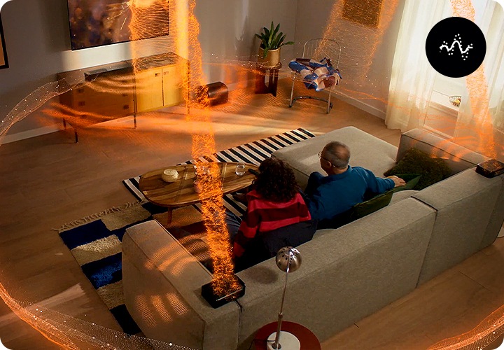 사람들이 소파에 앉아서 TV를 보고 있습니다. TV 아래에는 장식장이 놓여 있으며, 장식장 위에는 사운드바가 놓여 있고 장식장 오른쪽에는 우퍼가 놓여 있습니다. 소파 양 끝에 놓인 리어스피커와 사운드바, 우퍼에서 음향효과가 나오고 있습니다.