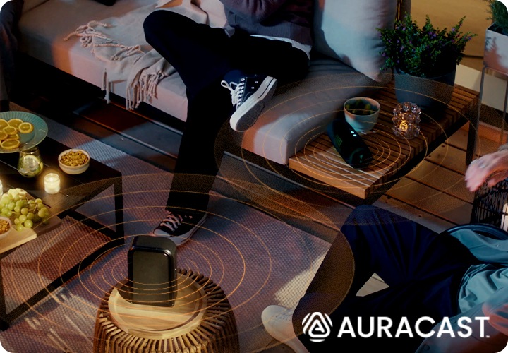 야외에 사람들이 소파에 앉아 있습니다. 테이블에는 리어스피커와 JBL 스피커가 놓여 있으며, 두개의 제품이 연결되어 있습니다.