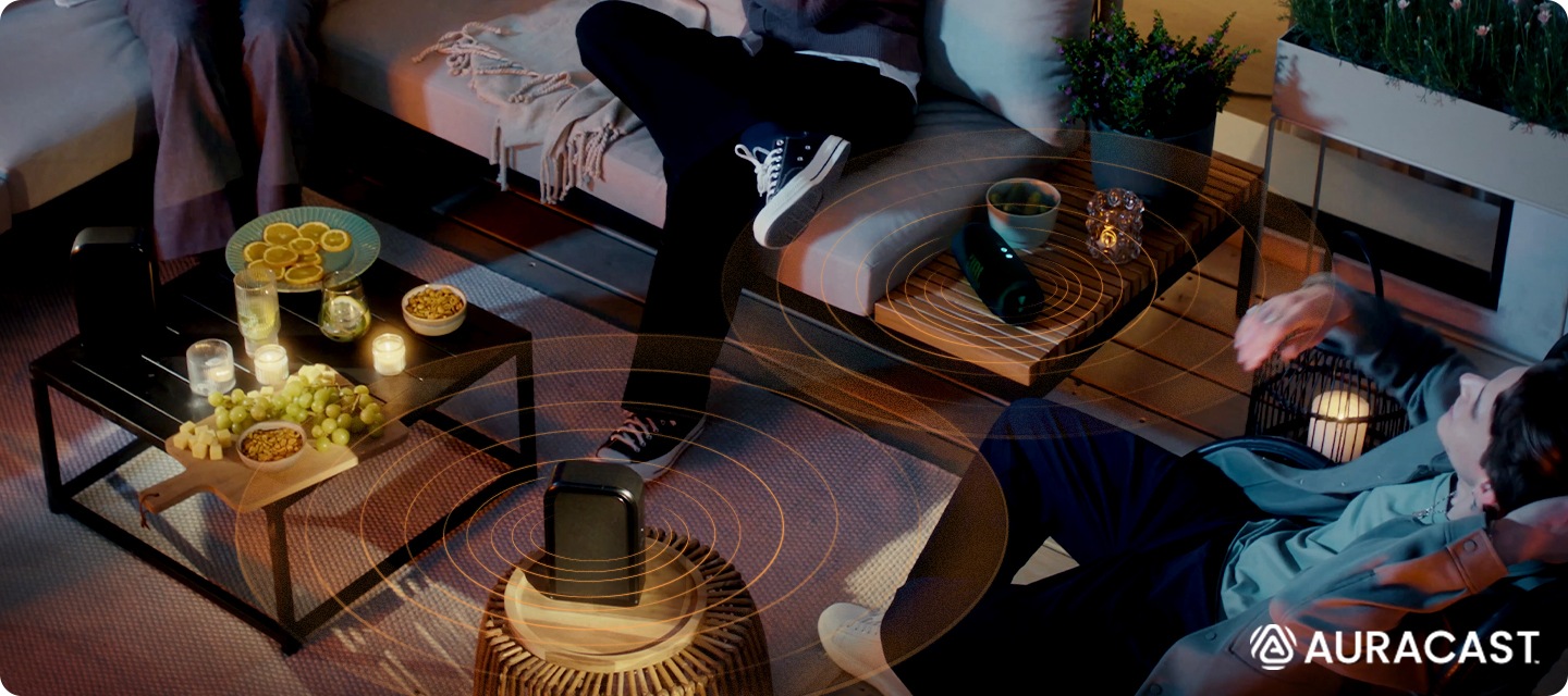 야외에 사람들이 소파에 앉아 있습니다. 테이블에는 리어스피커와 JBL 스피커가 놓여 있으며, 두개의 제품이 연결되어 있습니다.