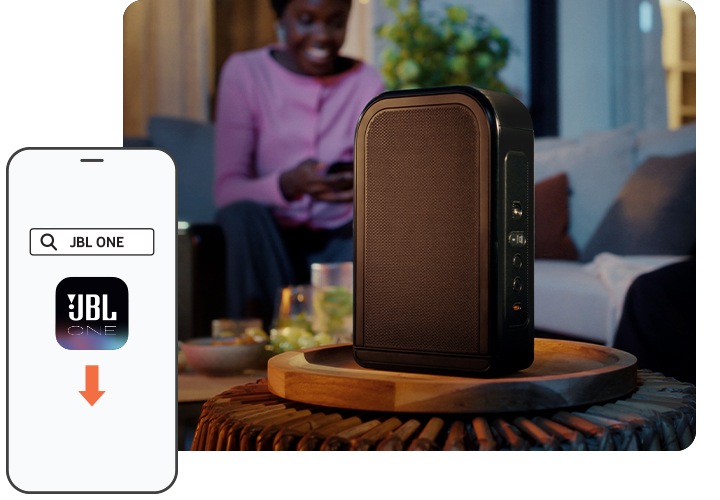 사람이 소파에 앉아 있습니다. 테이블에는 리어스피커가 놓여 있습니다. 이미지 왼쪽 하만에는 스마트폰이 보이며 화면 속에는 JBL ONE 앱의 아이콘이 보입니다.