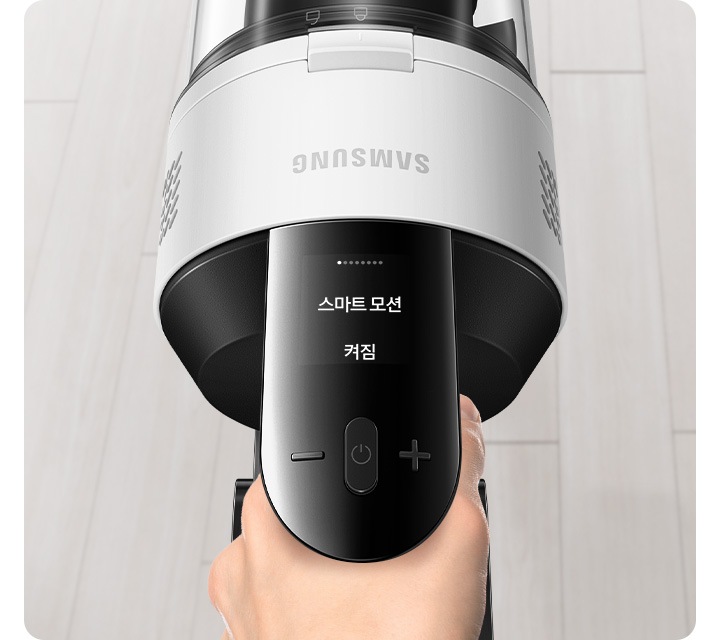 밝은 베이지 톤의 원목 마루가 깔린 바닥이 보입니다. 가운데는 제트 핏을 잡고 있는 손과 청소기 헤드가 확대되어 나타나 있고, 청소 중인 제트의 알림 창에는 스마트 모션 켜짐 문구가 보입니다.