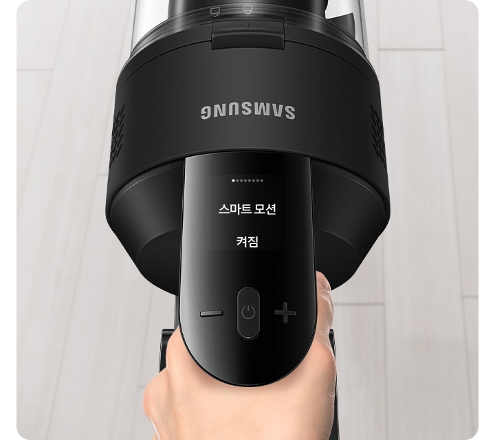 밝은 베이지 톤의 원목 마루가 깔린 바닥이 보입니다. 가운데는 제트 핏을 잡고 있는 손과 청소기 헤드가 확대되어 나타나 있고, 청소 중인 제트의 알림 창에는 스마트 모션 켜짐 문구가 보입니다.