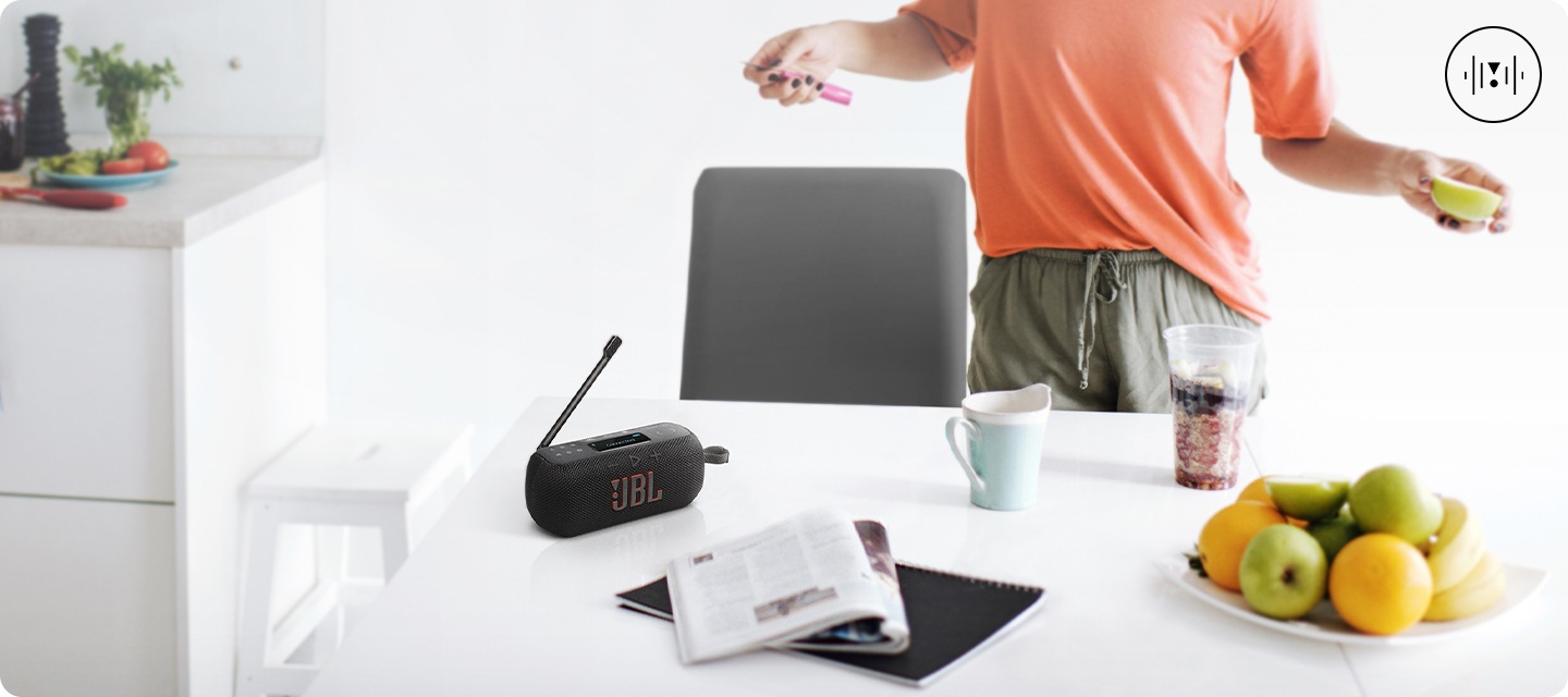 흰색 테이블 위에 블랙 색상의 라디오 스피커, 노트, 책, 컵들, 과일들이 놓여 있습니다. 테이블 뒤에는 사람이 서 있으며, 손에는 과도와 사과가 들려 있습니다. 