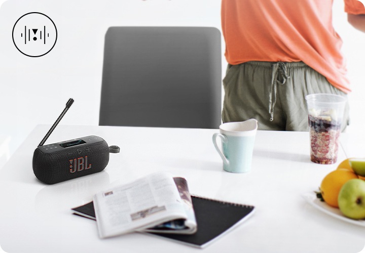 흰색 테이블 위에 블랙 색상의 라디오 스피커, 노트, 책, 컵들, 과일들이 놓여 있습니다. 테이블 뒤에는 사람이 서 있으며, 손에는 과도와 사과가 들려 있습니다. 