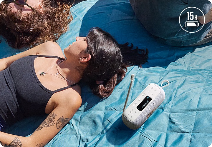 사람들이 하늘색 돗자리 위에 누워 있습니다. 어두운색 민소매를 입은 여성의 왼쪽에는 화이트 색상의 라디오 스피커가 놓여 있습니다.