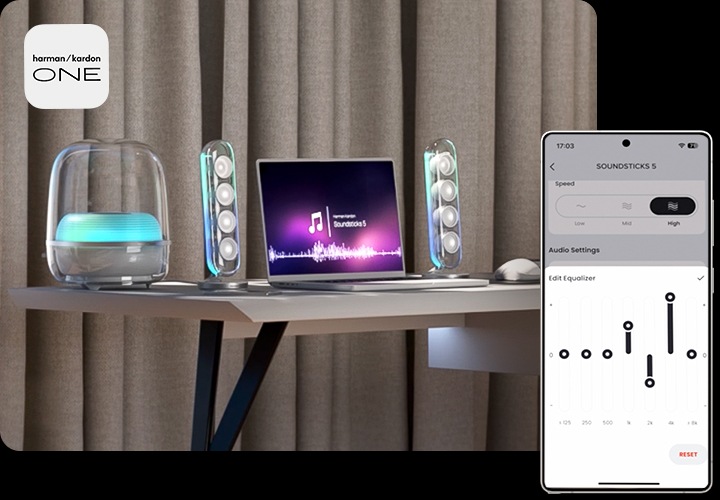 테이블 위에 노트북과 흰색 스피커가 놓여 있습니다. 노트북 화면에는 음악이 틀어졌는지 음표와 음향 효과가 나오고 있습니다. 이미지 오른쪽 하단으로는 휴대폰이 놓여 있으며, 화면에는 Harman Kardon One 앱 화면이 나오고 있습니다.