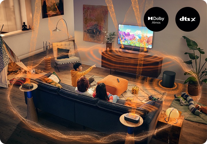 거실 벽에 TV와 사운드바가 설치되어 있으며, 사운드바 아래에는 장식장이 놓여 있습니다. 장식장 오른쪽 옆에는 서브우퍼가 놓여 있습니다. 장식장 앞에는 소파가 놓여 있으며, 소파에는 남성 한명과 여성 두명이 TV를 보고 있습니다. 사운드바와 서브우퍼에서는 음향효과가 나와 거실을 감싸고 있습니다.