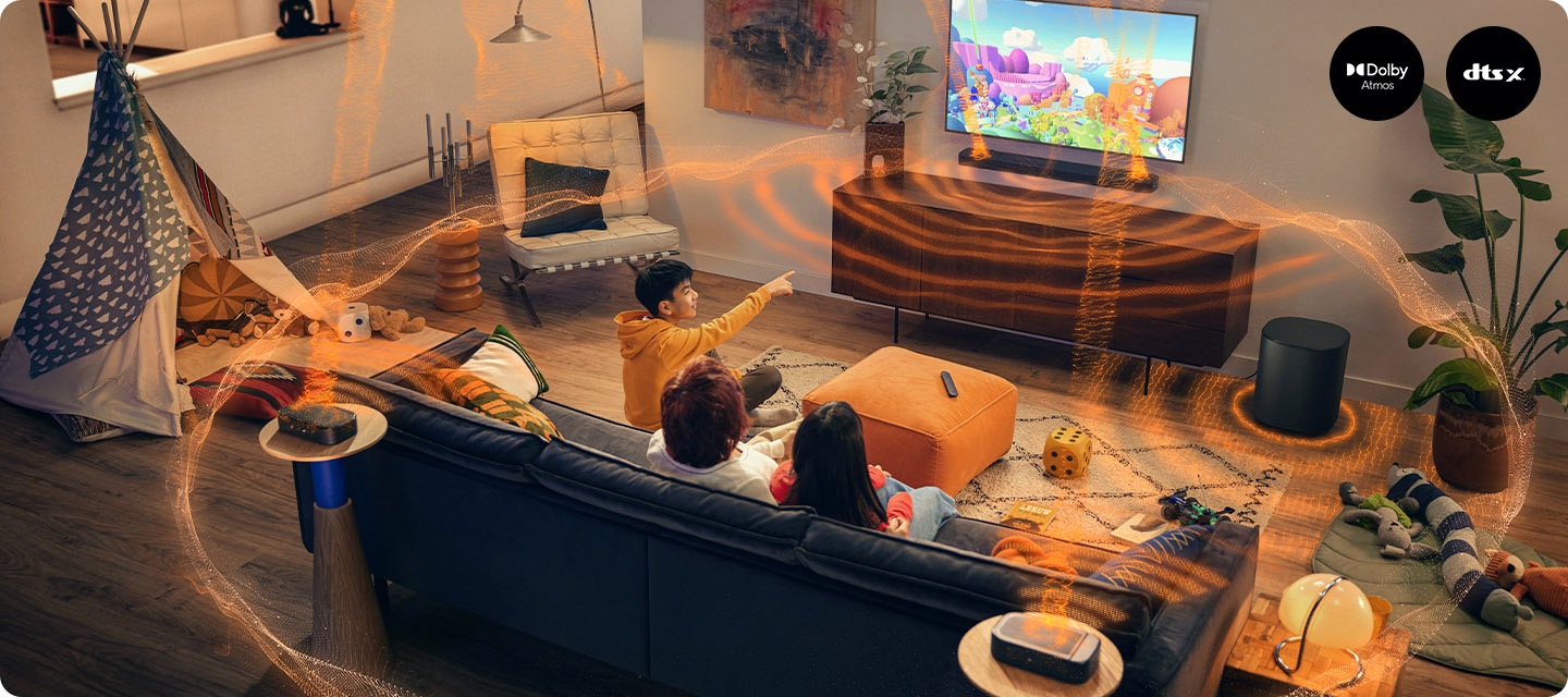 거실 벽에 TV와 사운드바가 설치되어 있으며, 사운드바 아래에는 장식장이 놓여 있습니다. 장식장 오른쪽 옆에는 서브우퍼가 놓여 있습니다. 장식장 앞에는 소파가 놓여 있으며, 소파에는 남성 한명과 여성 두명이 TV를 보고 있습니다. 사운드바와 서브우퍼에서는 음향효과가 나와 거실을 감싸고 있습니다.