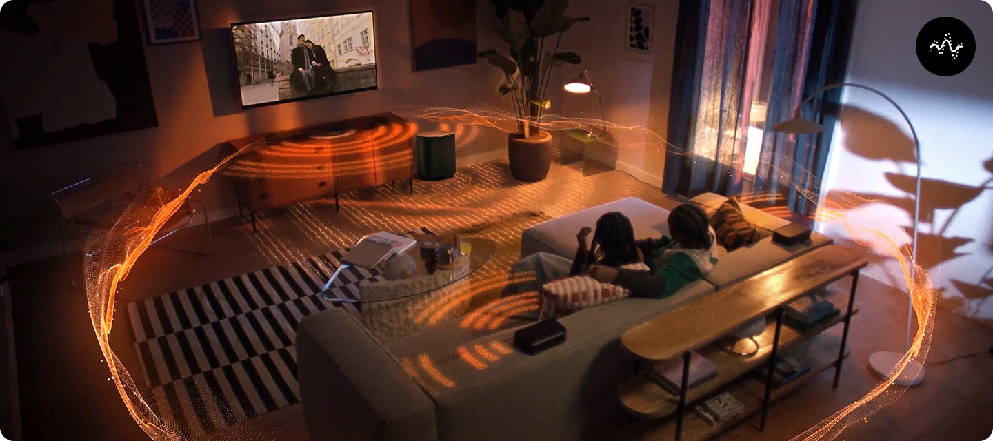 거실 벽에 TV와 사운드바가 설치되어 있으며, 사운드바 아래에는 장식장이 놓여 있습니다. 장식장 오른쪽 옆에는 서브우퍼가 놓여 있습니다. 장식장 앞에는 테이블과 소파가 놓여 있으며, 소파에는 남성 한명과 여성 한명이 TV를 보고 있습니다. 사운드바와 서브우퍼에서는 음향효과가 나와 거실을 감싸고 있습니다.