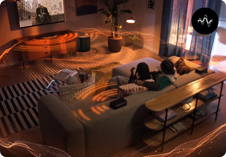 거실 벽에 TV와 사운드바가 설치되어 있으며, 사운드바 아래에는 장식장이 놓여 있습니다. 장식장 오른쪽 옆에는 서브우퍼가 놓여 있습니다. 장식장 앞에는 테이블과 소파가 놓여 있으며, 소파에는 남성 한명과 여성 한명이 TV를 보고 있습니다. 사운드바와 서브우퍼에서는 음향효과가 나와 거실을 감싸고 있습니다.