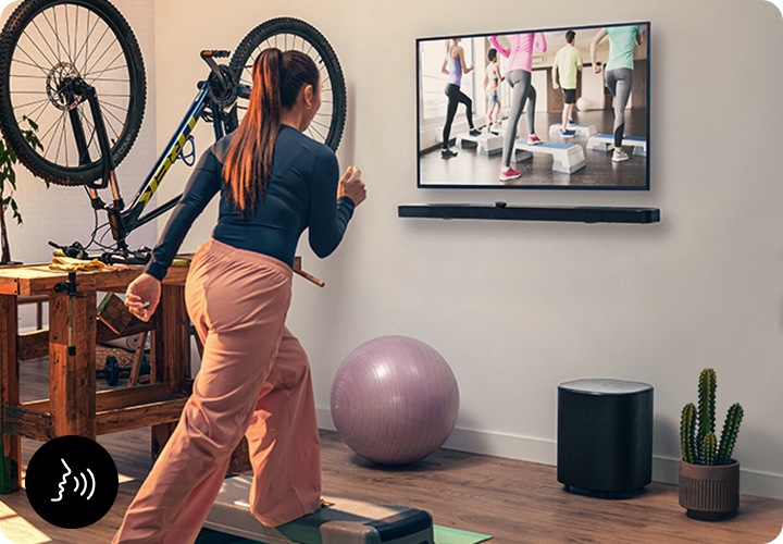 여성이 벽에 설치된 TV를 보며 운동을 하고 있습니다. TV 아래에는 사운드바가 설치되어 있으며, 바닥에는 서브우퍼가 놓여 있습니다.