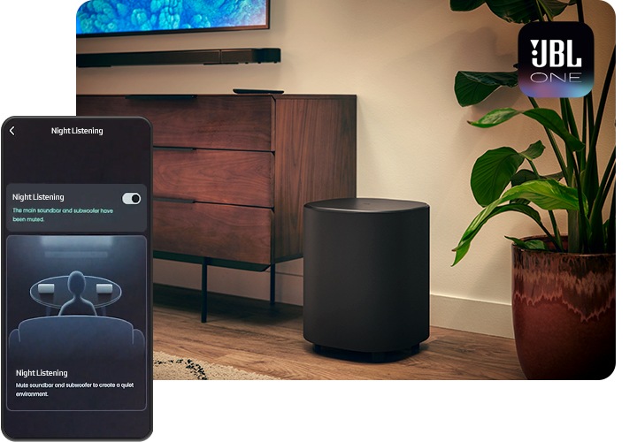 TV와 사운드바가 벽에 설치되어 있으며, 그 아래에는 장식장이 놓여 있습니다. 장식장 오른쪽 옆에는 서브우퍼와 화분이 놓여 있습니다. 이미지 왼쪽으로 휴대폰이 놓여 있으며, 스마트폰 화면에는 JBL ONE 앱 화면이 보입니다.