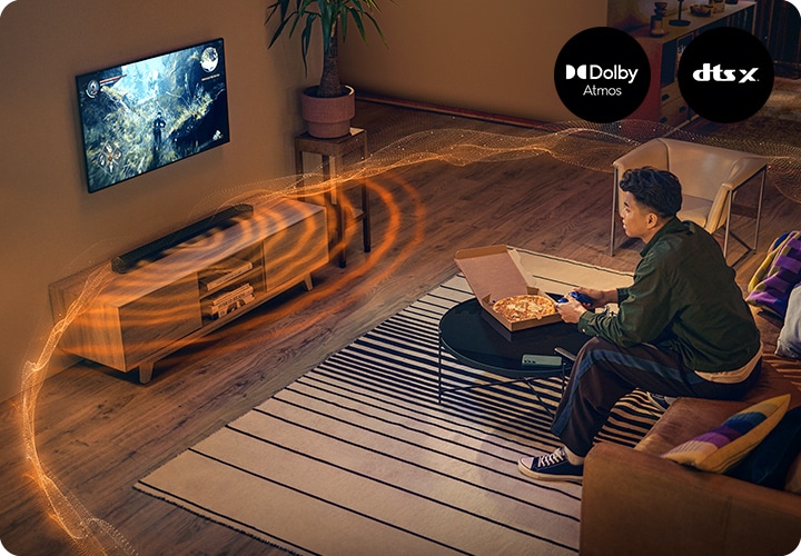 TV로 게임을 하는 남성이 소파에 앉아 있으며, 그 앞에는 피자가 놓여 있는 테이블이 있습니다. TV 아래에는 블랙 색상의 사운드바가 놓여 있으며, 사운드바에서는 음향효과가 나오고 있습니다.