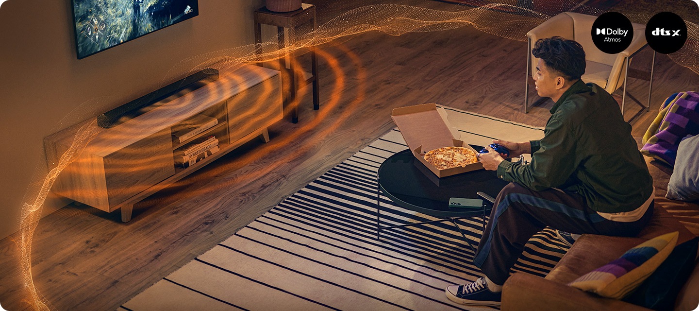 TV로 게임을 하는 남성이 소파에 앉아 있으며, 그 앞에는 피자가 놓여 있는 테이블이 있습니다. TV 아래에는 블랙 색상의 사운드바가 놓여 있으며, 사운드바에서는 음향효과가 나오고 있습니다.