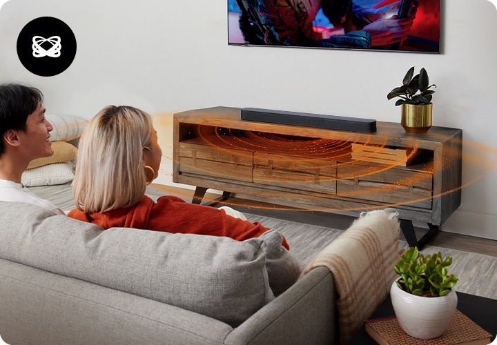 남성과 여성이 소파에 앉아서 벽에 설치된 TV를 시청하고 있습니다. TV 아래에는 장식장이 놓여 있으며, 그 위에는 블랙 색상의 사운드바가 놓여 있습니다. 사운드바에서는 음향효과가 나오고 있습니다.