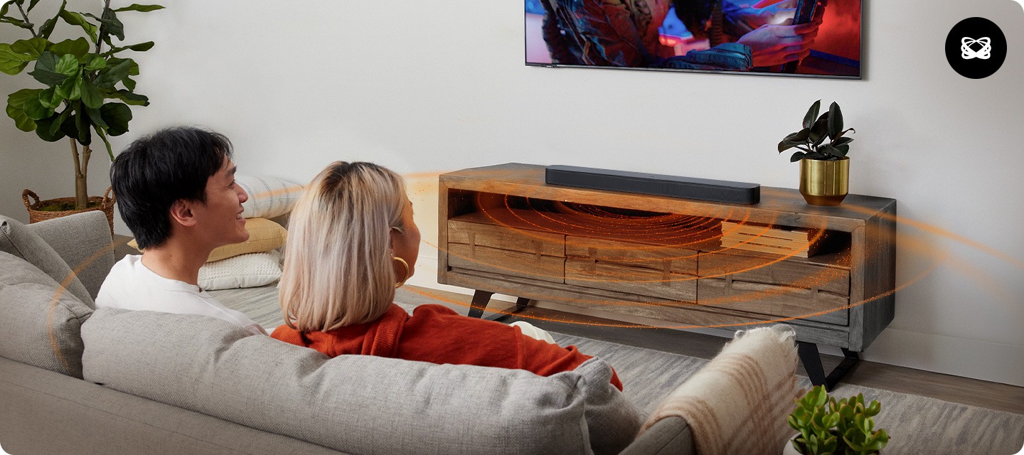 남성과 여성이 소파에 앉아서 벽에 설치된 TV를 시청하고 있습니다. TV 아래에는 장식장이 놓여 있으며, 그 위에는 블랙 색상의 사운드바가 놓여 있습니다. 사운드바에서는 음향효과가 나오고 있습니다.