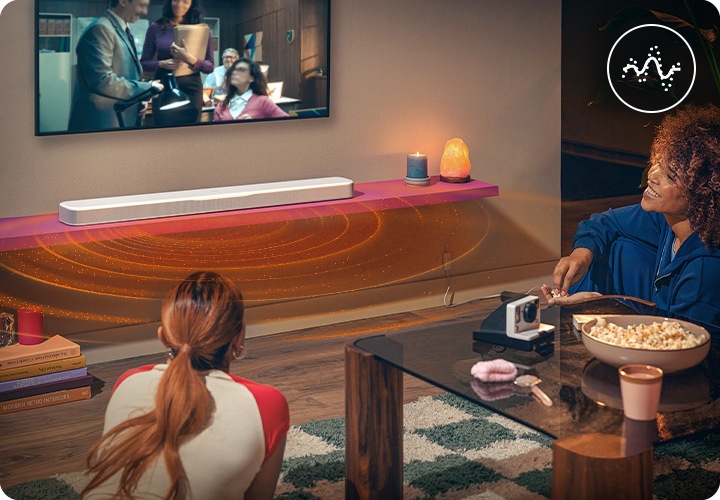 거실에서 여성 두명이 벽에 설치된 TV로 영화를 시청하고 있습니다. 여성들 사이에는 테이블이 놓여 있으며, 그 위에는 팝콘과 카메라가 놓여 있습니다. 긴 머리를 하나로 묶은 여성은 테이블 왼쪽에 엎드려 있으며, 곱슬머리를 한 여성은 테이블 오른쪽에 기대 앉아서 팝콘을 먹고 있습니다. TV 아래로는 선반이 설치되어 있으며, 그 위에 음향효과가 나오는 화이트 색상의 사운드바가 놓여 있습니다.