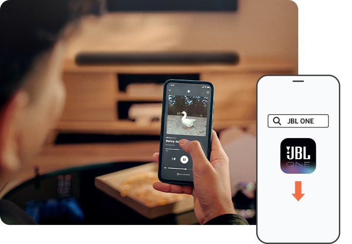 남성이 휴대폰으로 음악을 재생하고 있습니다. 남성 앞에는 블랙 색상의 사운드바가 놓여 있는 장식장이 보입니다. 이미지 오른쪽 하단에는 JBL One 앱 아이콘이 화면에 보이는 휴대폰이 놓여 있습니다.