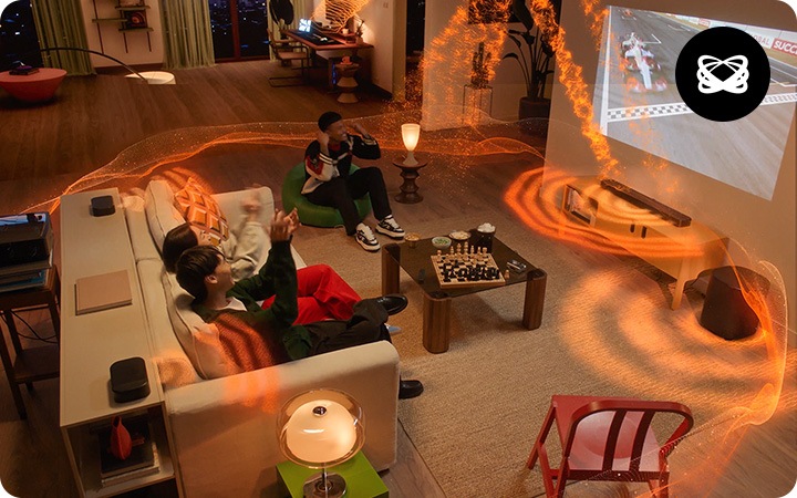 거실에 소파와 테이블 장식장들이 놓여 있습니다. 소파에는 여성과 남성이 2명이 앉아 있으며, 소파 왼쪽으로 녹색 1인용 소파가 남성 1명이 앉아 있습니다. 소파 앞으로는 테이블이 놓여 있으며 그 위에는 체스판이 놓여 있습니다. 소파 뒤에는 장식장이 놓여 있으며, 그 위에는 리어스피커가 놓여 있습니다. 장식장 뒤로는 빔 프로젝터가 놓여 있으며, 거실 벽에 레이싱 경기를 틀었습니다. 레이싱 경기가 나오는 화면 아래에는 사운드바가 놓인 장식장이 있습니다. 장식장 오른쪽에는 서브우퍼가 놓여 있습니다. 사운드바와 서브우퍼, 리어스피커에서 음향효과가 나옵니다.