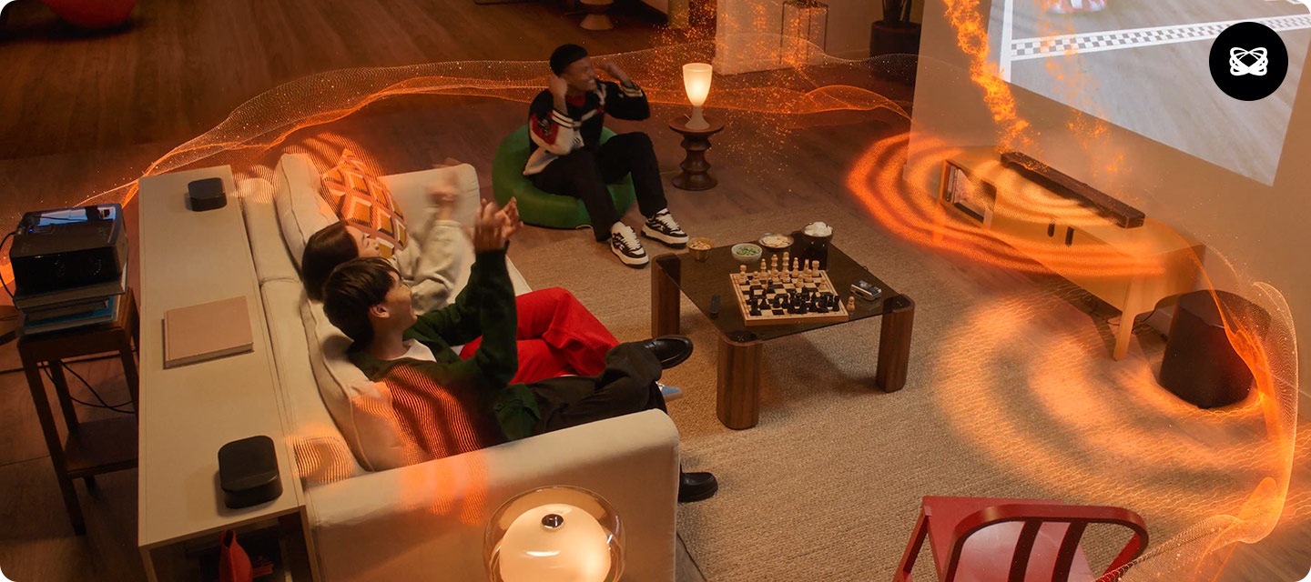 거실에 소파와 테이블 장식장들이 놓여 있습니다. 소파에는 여성과 남성이 2명이 앉아 있으며, 소파 왼쪽으로 녹색 1인용 소파가 남성 1명이 앉아 있습니다. 소파 앞으로는 테이블이 놓여 있으며 그 위에는 체스판이 놓여 있습니다. 소파 뒤에는 장식장이 놓여 있으며, 그 위에는 리어스피커가 놓여 있습니다. 장식장 뒤로는 빔 프로젝터가 놓여 있으며, 거실 벽에 레이싱 경기를 틀었습니다. 레이싱 경기가 나오는 화면 아래에는 사운드바가 놓인 장식장이 있습니다. 장식장 오른쪽에는 서브우퍼가 놓여 있습니다. 사운드바와 서브우퍼, 리어스피커에서 음향효과가 나옵니다.