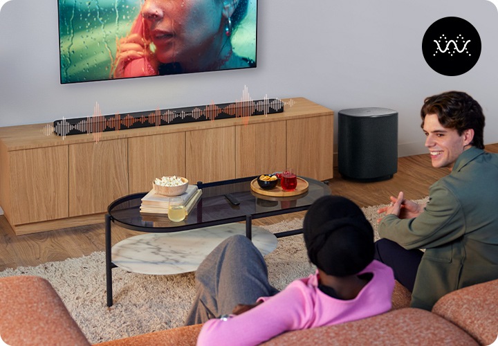 거실 벽에 TV가 설치되어 있으며, 그 아래에는 사운드바가 놓인 장식장이 있습니다. 장식장 오른쪽에는 서브우퍼가 놓여 있습니다. 장식장 앞에는 테이블과 소파가 있으며, 소파에는 2명의 사람이 앉아 있습니다. 사운드바에는 음향효과가 표시되어 있습니다.