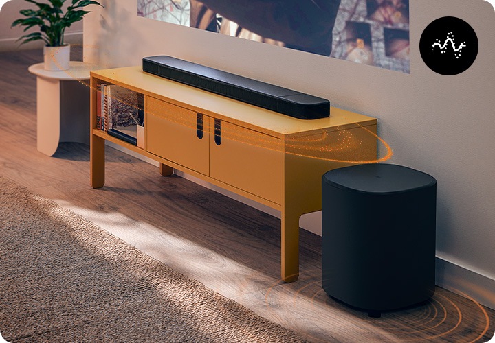 사운드바가 놓인 장식장 위에는 영화의 한 장면이 나오고 있습니다. 장식장 오른쪽에는 서브우퍼가 놓여 있으며, 왼쪽에는 화분이 놓인 조금한 테이블이 있습니다. 사운드바 양 끝에 달린 리어스피커에서 음향효과가 나오고 있습니다.