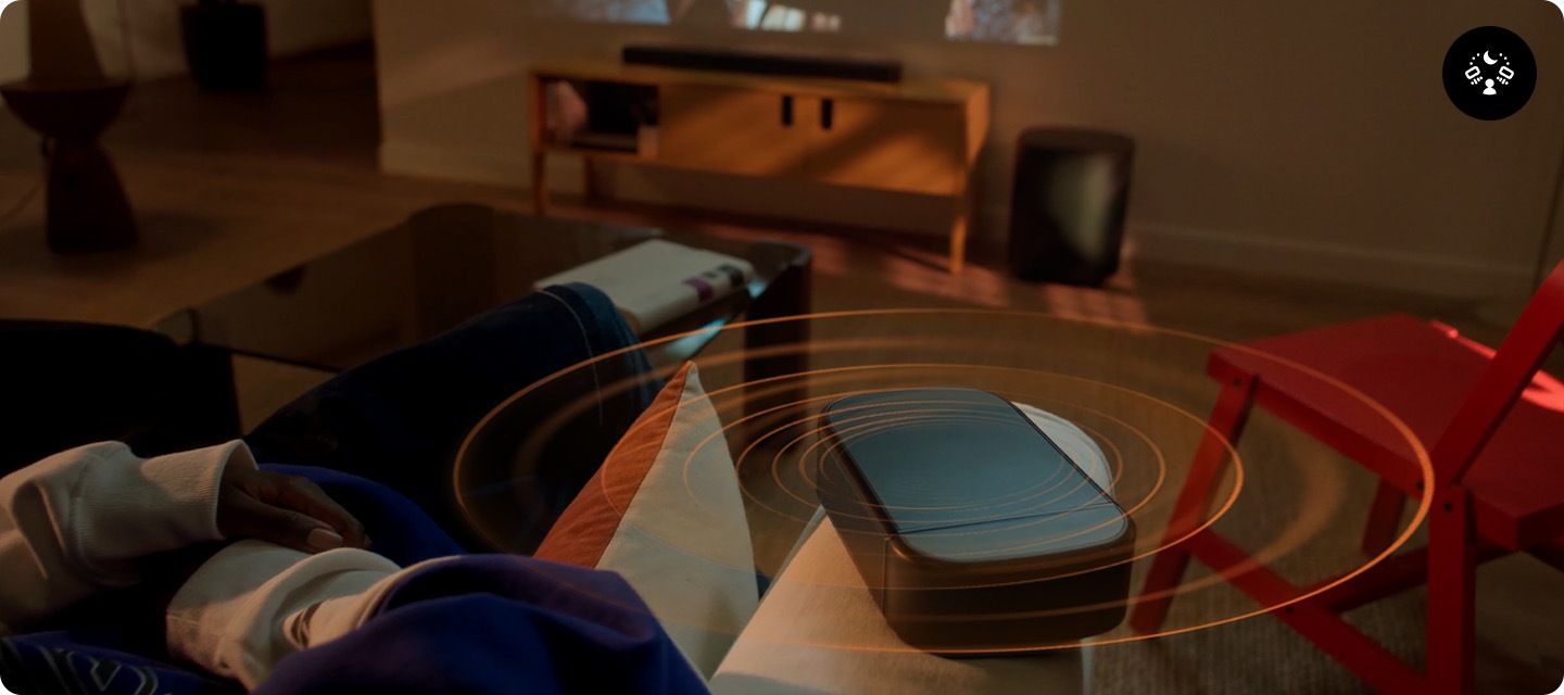 사운드바가 놓인 장식장 위에는 영화의 한 장면이 나오고 있습니다. 장식장 오른쪽에는 서브우퍼가 놓여 있습니다. 장식장 앞에는 소파에 앉아서 영화를 보는 사람이 있으며, 소파 손잡이 위에는 리어스피커가 놓여 있습니다. 사운드바와 서브우퍼, 리어스피커에서는 음향효과가 나오고 있습니다.