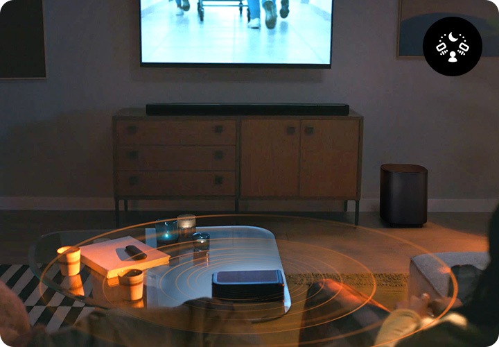 거실 벽에 TV가 설치되어 있으며, TV 아래에는 장식장이 놓여 있습니다. 장식장 위에는 사운드바가 놓여 있고 장식장 오른쪽 옆에는 서브우퍼가 놓여 있습니다. 장식장 앞에는 테이블과 소파가 놓여 있으며, 소파에는 한 사람은 누워 있고, 한사람은 앉아서 TV를 보고 있습니다. 테이블 위에는 책과 컵들 그리고 리어 스피커가 놓여 있으며 사운드바와 리어 스피커, 서브우퍼에서는 음향효과가 나옵니다