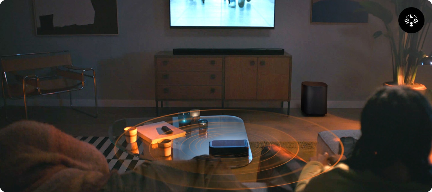 거실 벽에 TV가 설치되어 있으며, TV 아래에는 장식장이 놓여 있습니다. 장식장 위에는 사운드바가 놓여 있고 장식장 오른쪽 옆에는 서브우퍼가 놓여 있습니다. 장식장 앞에는 테이블과 소파가 놓여 있으며, 소파에는 한 사람은 누워 있고, 한사람은 앉아서 TV를 보고 있습니다. 테이블 위에는 책과 컵들 그리고 리어 스피커가 놓여 있으며 사운드바와 리어 스피커, 서브우퍼에서는 음향효과가 나옵니다