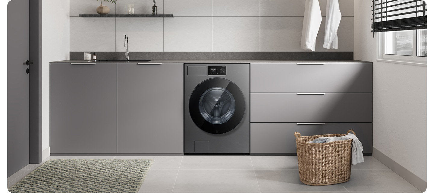 화이트&그레이톤 인테리어컷으로 그레이톤 하부장 중앙에 Bespoke AI 세탁기가 설치되어 있습니다. 상부장 부분에는 싱크대와 수전이 있고 앞쪽에는 라탄 바구니에 옷감이 들어있는 연출된 이미지입니다.