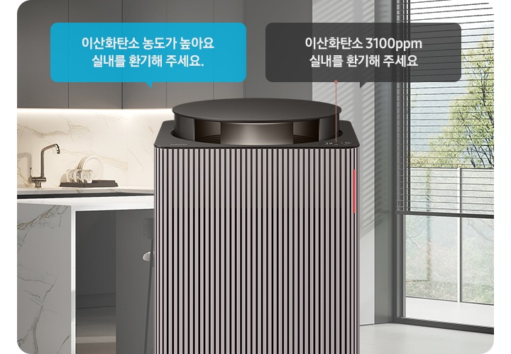그레이 톤의 주방 전경을 배경으로 오른쪽에는 아일랜드 식탁 의자 2개와 블라인드가 내려진 커다란 창문이 놓여 있습니다. 이미지 가운데에 Infinite AI 공기청정기 상단부가 확대되어 보입니다. 공기청정기 상단에는 알림 창 부분이 이산화탄소 3100ppm 실내를 환기해 주세요. 가 확대되어 놓여 있고 공기청정기 오른쪽으로 음성 안내를 표시하는 파란색 말풍선 안에 이산화탄소 농도가 높아요 실내를 환기해 주세요.라는 문구가 적혀 있습니다.