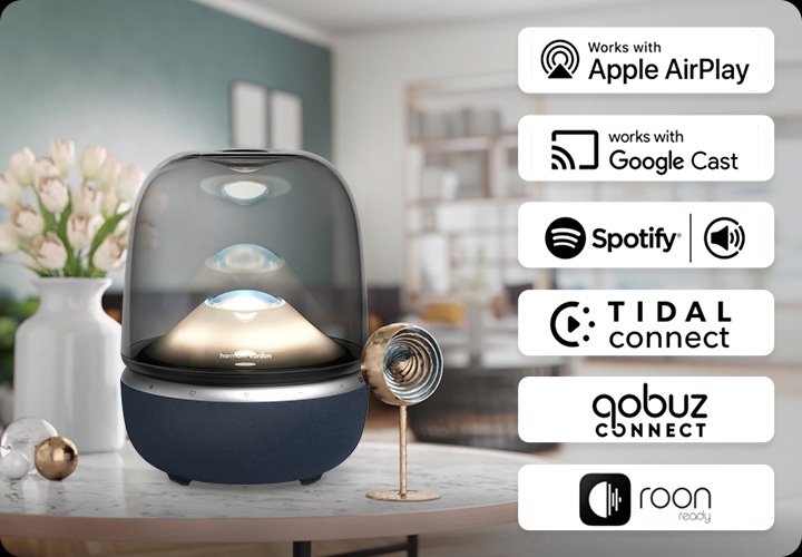 거실 테이블 위에 스피커가 놓여 있습니다. 스피커 오른쪽에는 AirPlay, Google Cast, Spotify Connect, Tidal Connec, Qobuz Connect, Roon ready의 아이콘이 있습니다.