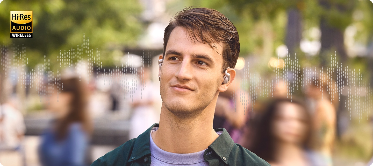 그레이 색상의 이어폰을 착용한 남성이 사람들이 걸어 다니고 있는 길거리에 서 있습니다. 길을 걸어 다니는 사람들은 흐릿하게 보입니다. 남성의 양 옆으로는 음향효과가 보입니다.