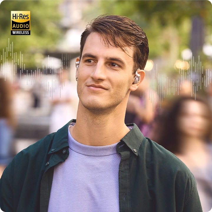 그레이 색상의 이어폰을 착용한 남성이 사람들이 걸어 다니고 있는 길거리에 서 있습니다. 길을 걸어 다니는 사람들은 흐릿하게 보입니다. 남성의 양 옆으로는 음향효과가 보입니다.