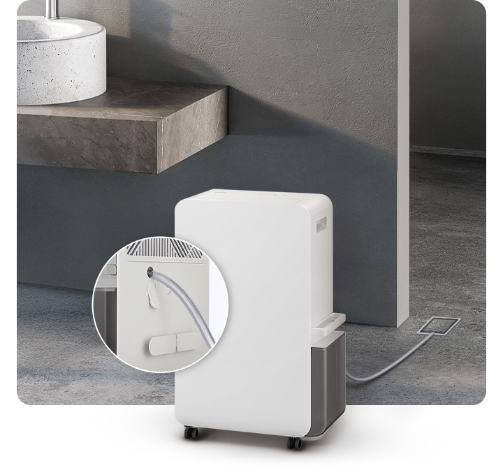 진한 회색의 화장실을 배경으로 이미지 왼쪽 부분에는 화이트 색상의 대리석으로 된 세면대와 브라운이 약간 섞인 색상의 세면대 받침대가 자리하고 있으며, 이미지 중간 하단 부분에는 오른쪽 측면이 약간 보이는 제습기의 모습이 있습니다. 제습기 뒤로 배수 호스가 하수구와 연결되어 있으며, 제습기 왼쪽 부분에는 배수 호스가 제습기와 연결되는 부분을 확대하여 자세히 알려주고 있습니다.