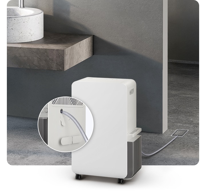 진한 회색의 화장실을 배경으로 이미지 왼쪽 부분에는 화이트 색상의 대리석으로 된 세면대와 브라운이 약간 섞인 색상의 세면대 받침대가 자리하고 있으며, 이미지 중간 하단 부분에는 오른쪽 측면이 약간 보이는 제습기의 모습이 있습니다. 제습기 뒤로 배수 호스가 하수구와 연결되어 있으며, 제습기 왼쪽 부분에는 배수 호스가 제습기와 연결되는 부분을 확대하여 자세히 알려주고 있습니다.