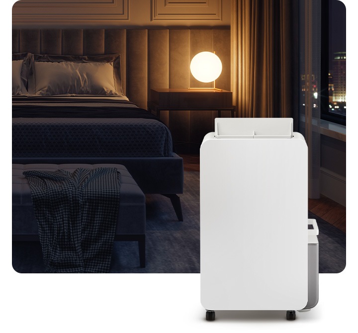 불이 꺼진 침실을 배경으로 침대 옆 원형 조명 하나가 켜져있고 조명 오른쪽 부분에는 작게 야경이 보이는 창이 있습니다. 이미지 오른쪽 하단 부분에 작동 중인 제습기가 서 있습니다.