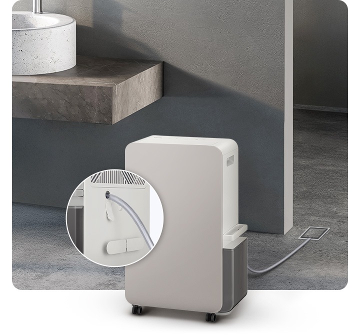 진한 회색의 화장실을 배경으로 이미지 왼쪽 부분에는 화이트 색상의 대리석으로 된 세면대와 브라운이 약간 섞인 색상의 세면대 받침대가 자리하고 있으며, 이미지 중간 하단 부분에는 오른쪽 측면이 약간 보이는 제습기의 모습이 있습니다. 제습기 뒤로 배수 호스가 하수구와 연결되어 있으며, 제습기 왼쪽 부분에는 배수 호스가 제습기와 연결되는 부분을 확대하여 자세히 알려주고 있습니다.