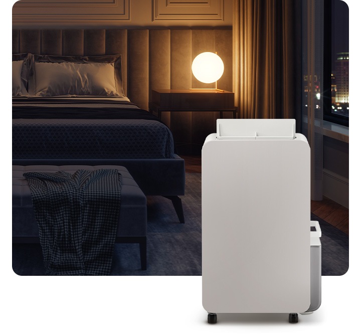 불이 꺼진 침실을 배경으로 침대 옆 원형 조명 하나가 켜져있고 조명 오른쪽 부분에는 작게 야경이 보이는 창이 있습니다. 이미지 오른쪽 하단 부분에 작동 중인 제습기가 서 있습니다.