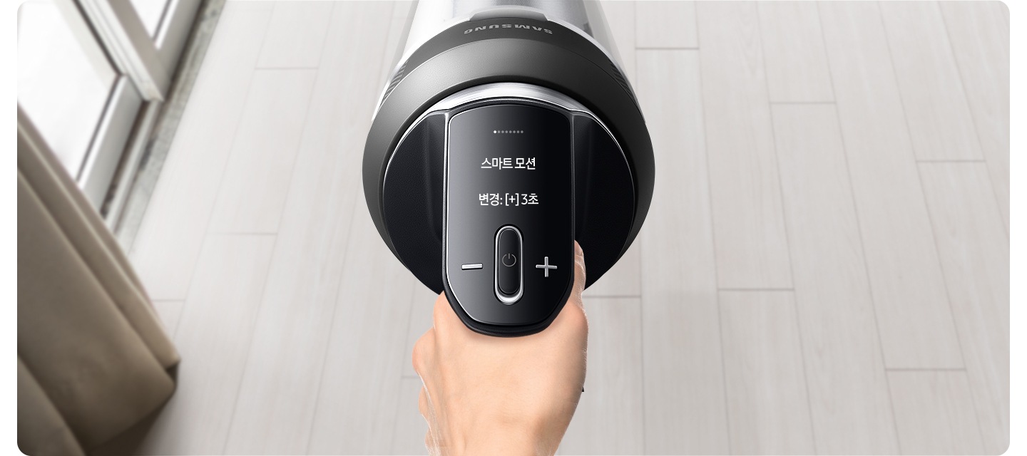 밝은 베이지 톤의 원목 마루가 깔린 바닥을 배경으로 왼쪽에는 베란다 창틀이 위치하고 있고, 그 옆으로 갈색 톤의 커튼 자락이 함께 위치하여 있습니다. 이미지 가운데 부분에는 제트를 잡고 있는 손과 대화형 알림창을 확대하여 상태를 알려주고 있습니다. 확대된 LCD 화면에는 스마트 모션 변경 : [+]3초가 쓰여 있습니다.