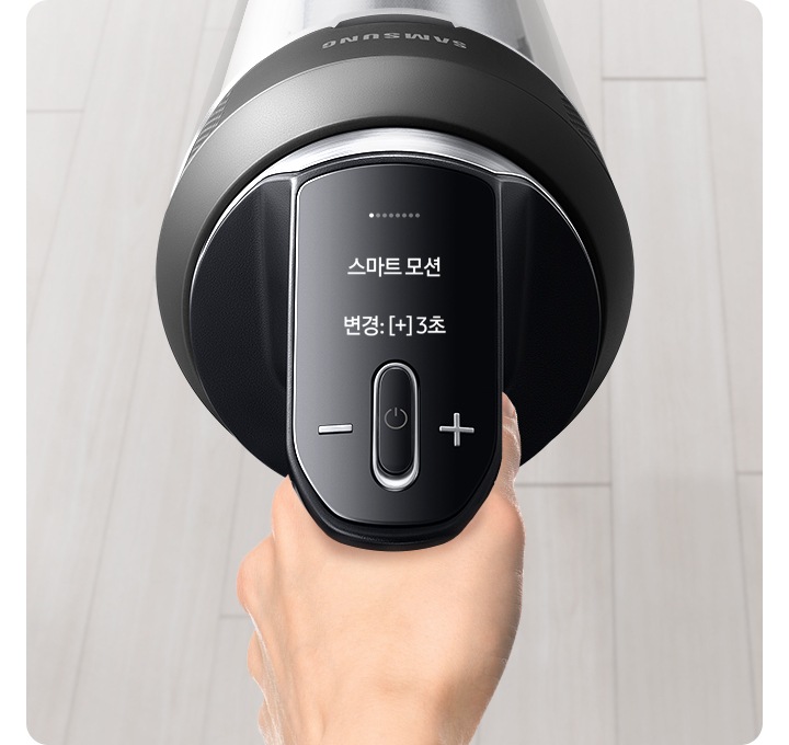 밝은 베이지 톤의 원목 마루가 깔린 바닥을 배경으로 왼쪽에는 베란다 창틀이 위치하고 있고, 그 옆으로 갈색 톤의 커튼 자락이 함께 위치하여 있습니다. 이미지 가운데 부분에는 제트를 잡고 있는 손과 대화형 알림창을 확대하여 상태를 알려주고 있습니다. 확대된 LCD 화면에는 스마트 모션 변경 : [+]3초가 쓰여 있습니다.