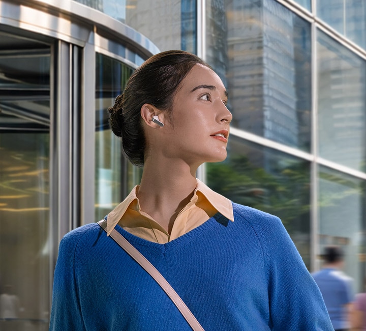 빌딩 앞에 한쪽 귀에 화이트 색상의 갤럭시 버즈4 이어버드를 착용한 여성이 오른쪽으로 고개를 향하고 있습니다.